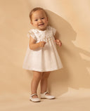 Vestido de bebé blanco en batista bordada con cuello volante y lazos en hombros, zapatos blancos.
