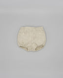 Culotte bebé de lino rústico beige con bolsillos de ojal y gomas en pierna, vista trasera sobre fondo gris.