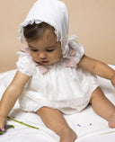 Bebé con vestido blanco de tul bordado y encaje, lazos rosas en hombros y capota a juego para bautizo