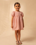 Vestido de lino rosa para niña con manga farol y falda amplia, combinado con sandalias grises.